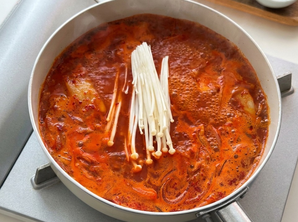 끓고 있는 붉은 김치찌개 팬에 생 팽이버섯 한 줌을 넣는 모습.