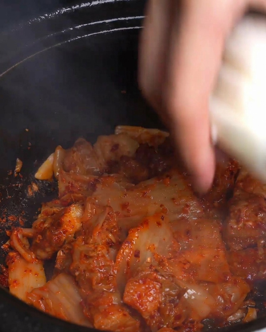 새빨갛게 볶아진 김치와 돼지고기가 담긴 뜨거운 냄비에 두툼하게 썬 양파를 넣는 모습.