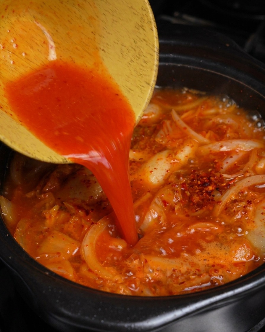 나무 그릇에 담긴 붉은 김칫국물을 끓고 있는 김치찌개에 붓는 모습.