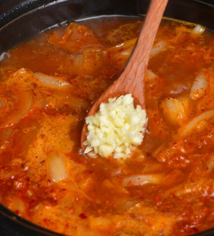 나무 숟가락으로 끓고 있는 빨간 김치찌개에 다진 마늘을 넣는 모습.