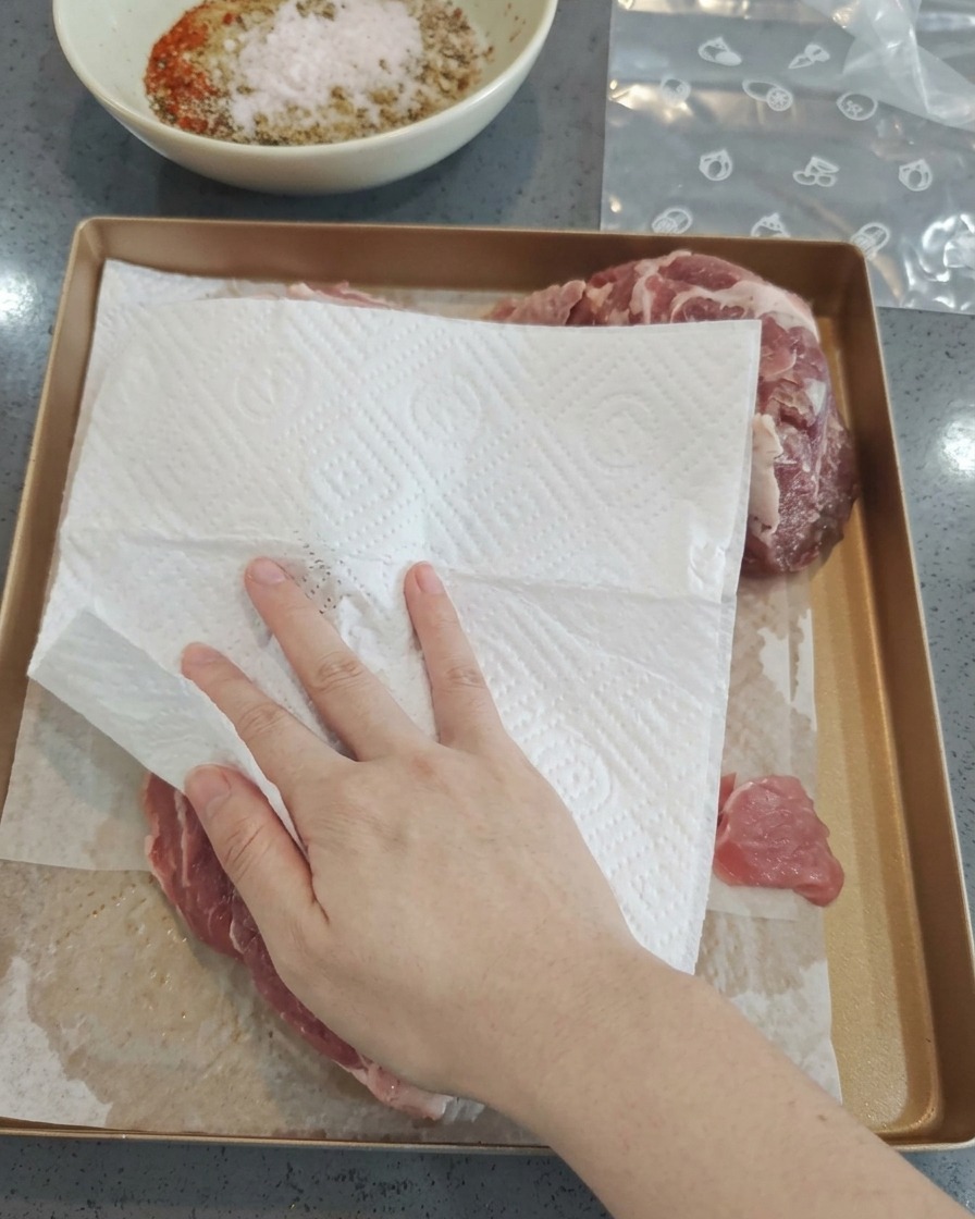 쟁반 위 돼지고기를 흰색 키친타월로 닦아내어 수분을 제거하는 모습