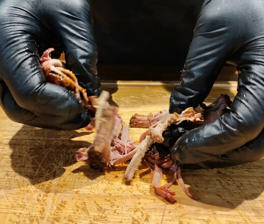 나무 도마 위에서 장갑을 낀 손으로 부드럽게 익은 이베리코 돼지 목살을 결대로 찢고 있는 모습