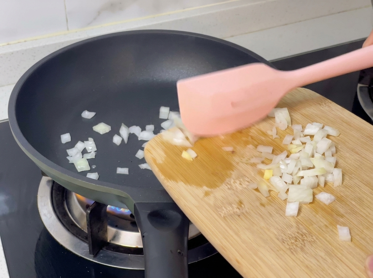 도마 위의 다진 양파를 분홍색 주걱으로 뜨거운 팬에 옮겨 담는 모습.