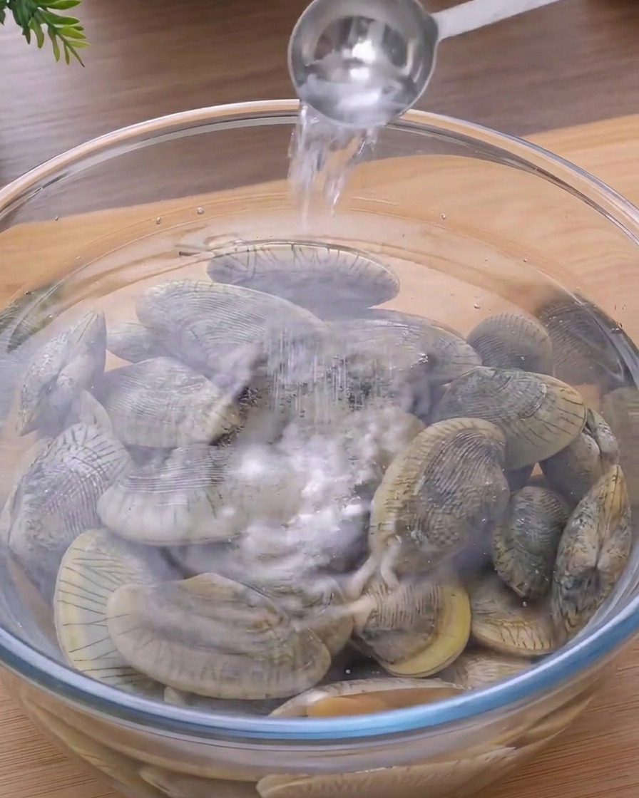 물과 신선한 조개가 담긴 투명한 유리 볼에 숟가락으로 소금을 넣는 모습.