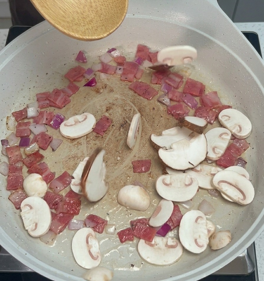 베이컨과 양파가 담긴 팬에 슬라이스한 버섯들을 넣는 모습.