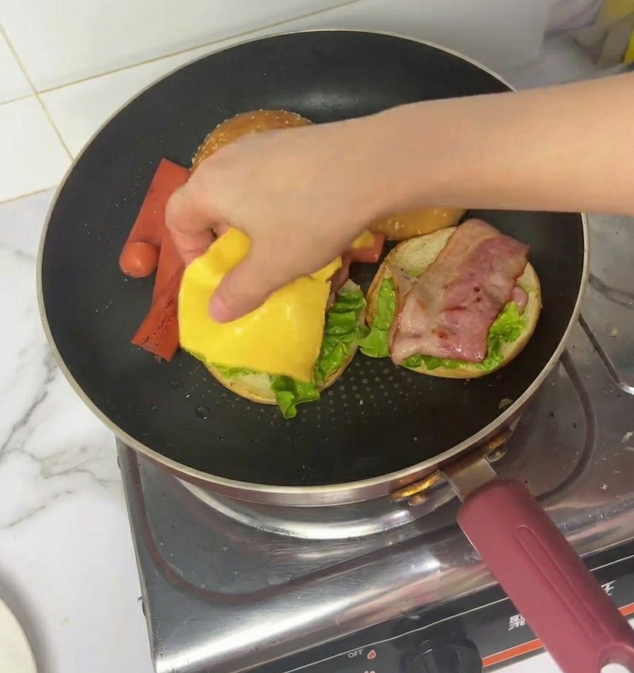 프라이팬 안에서 상추와 베이컨이 올라간 버거 위에 노란색 사각 치즈를 올리는 손.