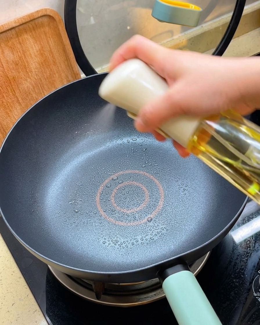 가열된 검은색 논스틱 팬에 오일 스프레이로 기름을 얇게 뿌리는 모습.