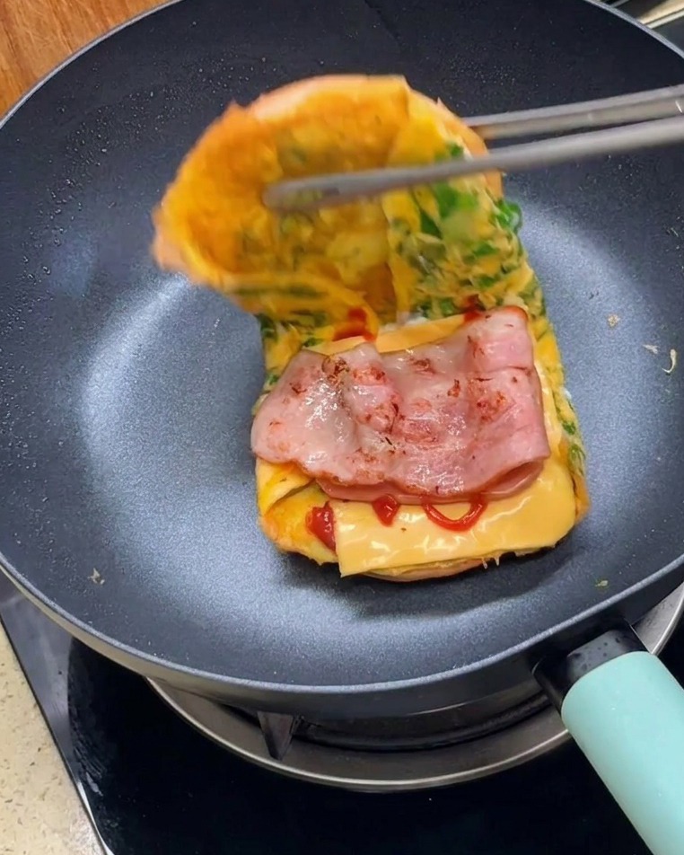 집게를 사용하여 계란으로 감싼 버거 번의 양쪽을 겹쳐 샌드위치를 완성하는 모습.