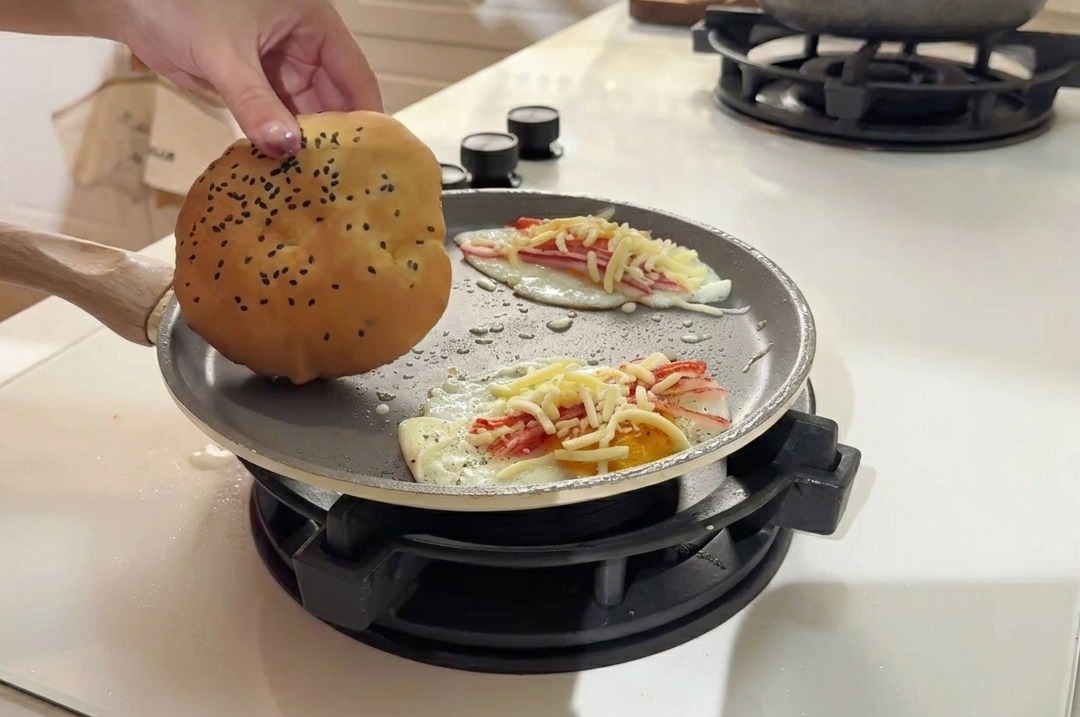 계란 패티 옆 팬 빈 공간에 참깨가 뿌려진 황금빛 버거 번을 올려두는 모습.