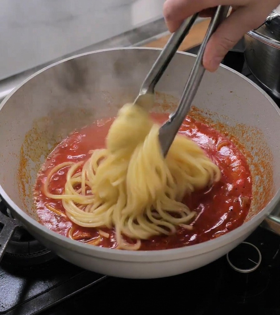 집게를 사용하여 삶아진 부카티니 면을 냄비에서 토마토 소스가 끓는 팬으로 바로 옮기는 모습.