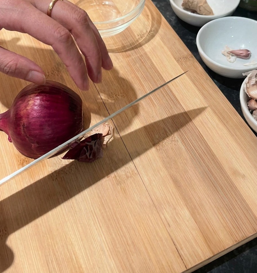 도마 위에서 적양파의 양 끝을 자르고 껍질을 벗긴 뒤 다지기 위해 칼을 든 손.