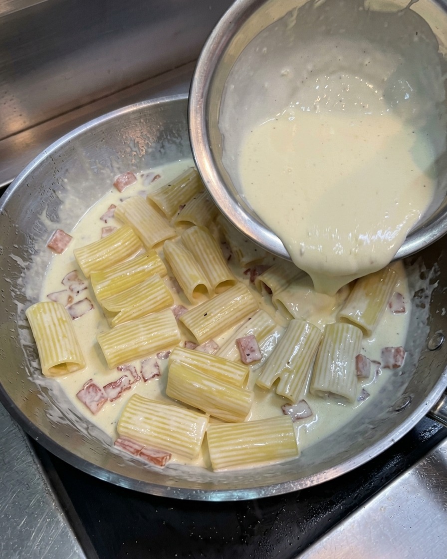 팬 안의 파스타 위에 노란 계란과 치즈 혼합물을 붓는 모습.