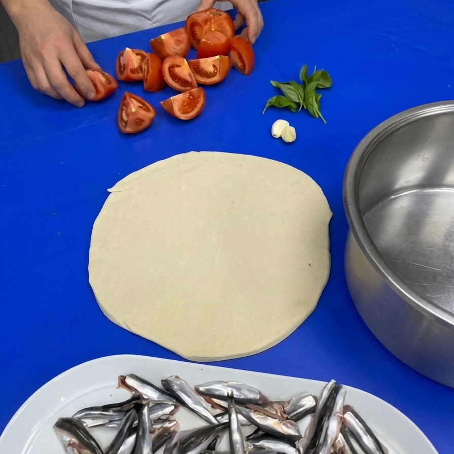 파란색 작업대 위에서 요리사가 4등분한 토마토, 마늘 한 쪽, 신선한 바질 잎을 집어 들고 있는 모습