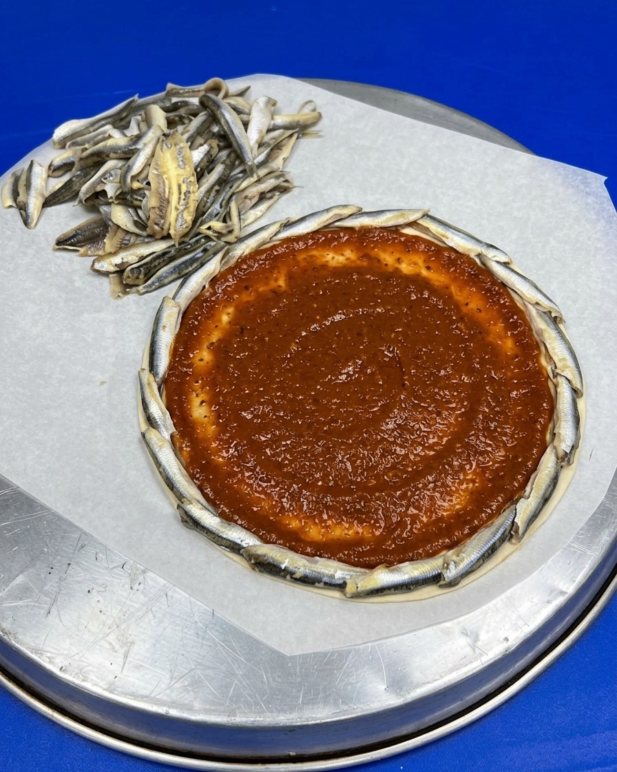 멸치 테두리가 둘러진 원형 도우 중앙에 붉은 토마토 소스가 매끄럽게 발려 있는 사진