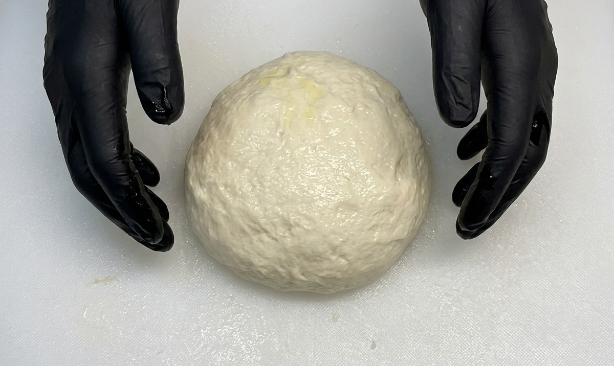 검은 장갑을 낀 손으로 흰 도마 위에서 매끄럽고 둥근 피자 반죽 공을 성형하는 모습.