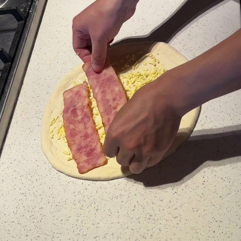 치즈와 소스를 올린 피자 위에 데친 베이컨 조각을 넓게 배치하는 모습.