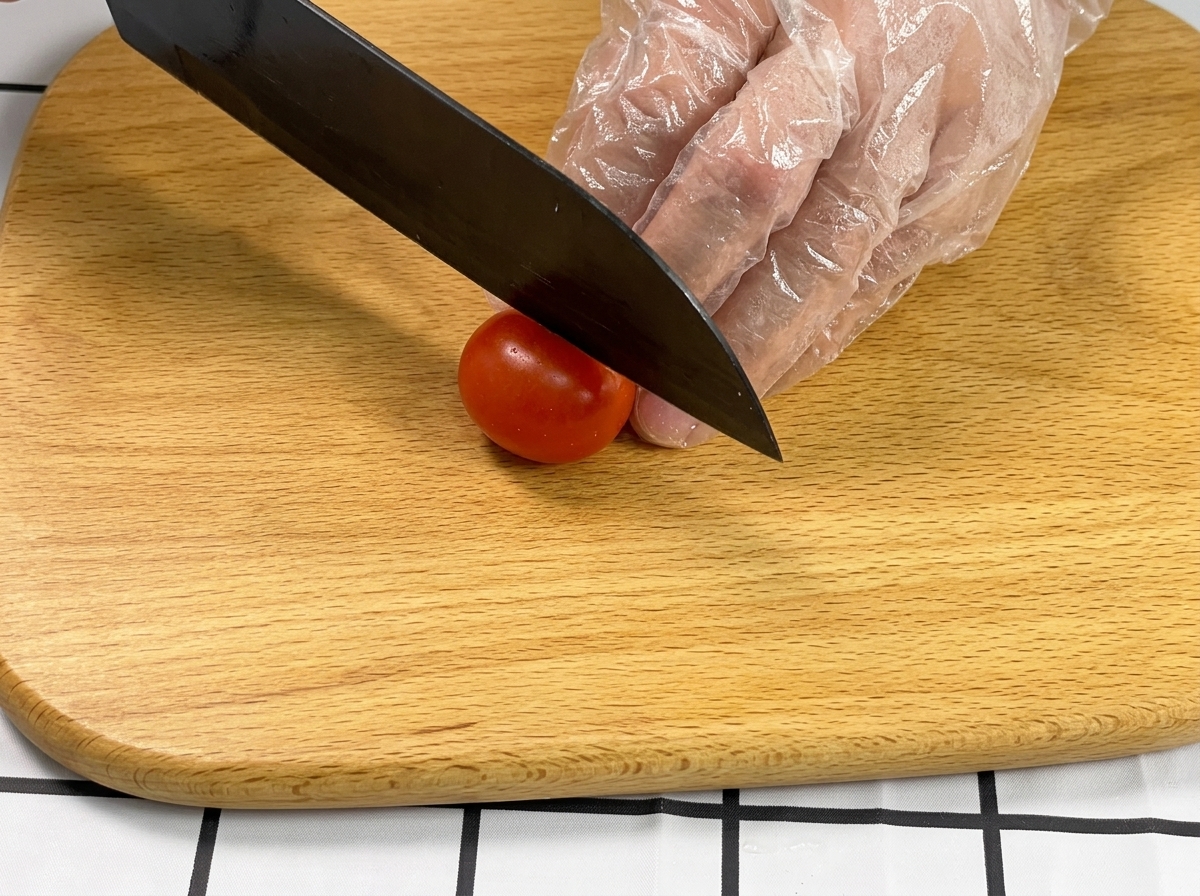 장갑 낀 손으로 방울토마토를 잡고 나무 도마 위에서 검은색 칼로 반을 자르는 모습.