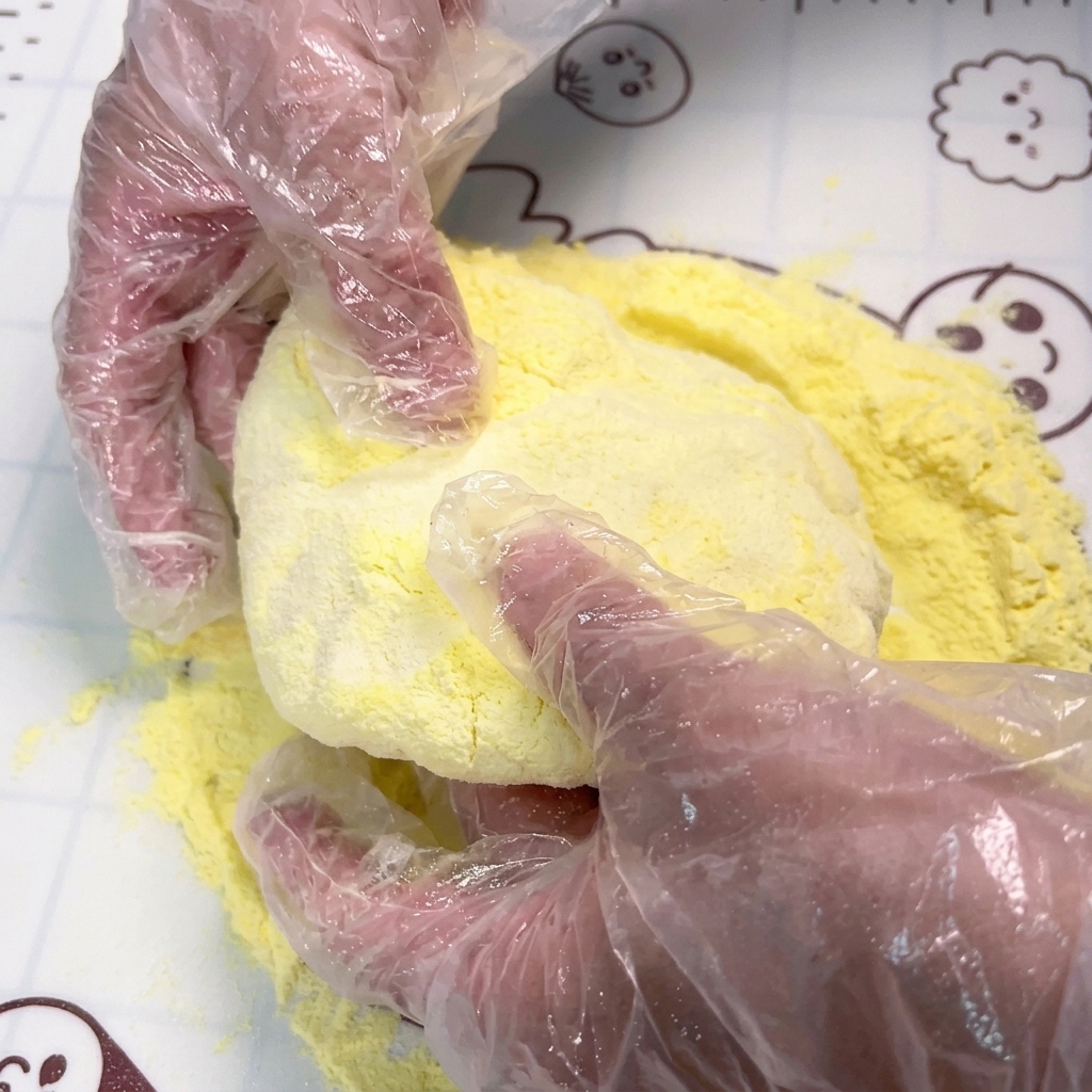 노란 옥수수가루가 뿌려진 바닥 위에서 장갑 낀 손으로 둥근 반죽을 누르며 펴는 모습.