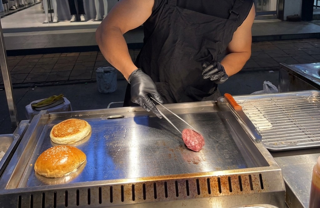 집게를 사용하여 동그랗게 뭉친 소고기 반죽을 뜨거운 금속 그릴 위에 올리는 모습.