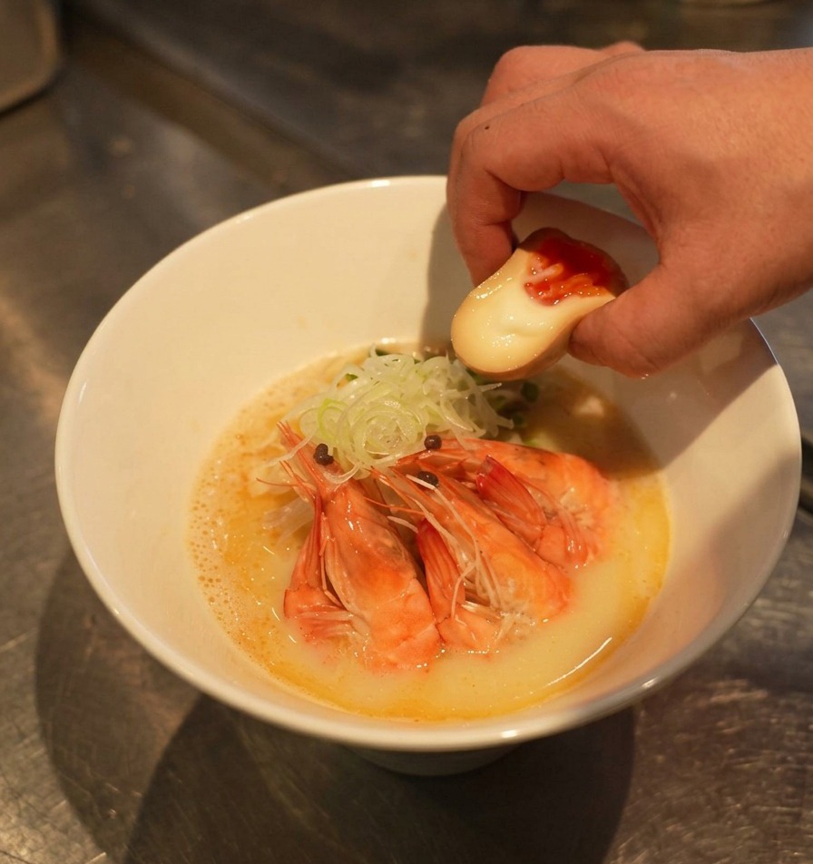 새우와 대파가 올라간 라멘 그릇에 노른자가 반짝이는 반숙 맛달걀 반 알을 곁들이는 모습.