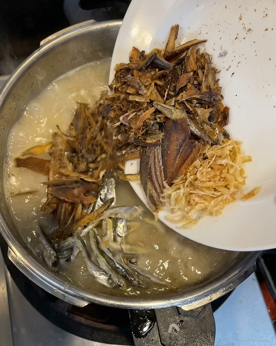 흰 접시에 담긴 말린 생선 가루, 은빛 멸치, 건새우를 부드럽게 끓고 있는 육수에 넣는 모습