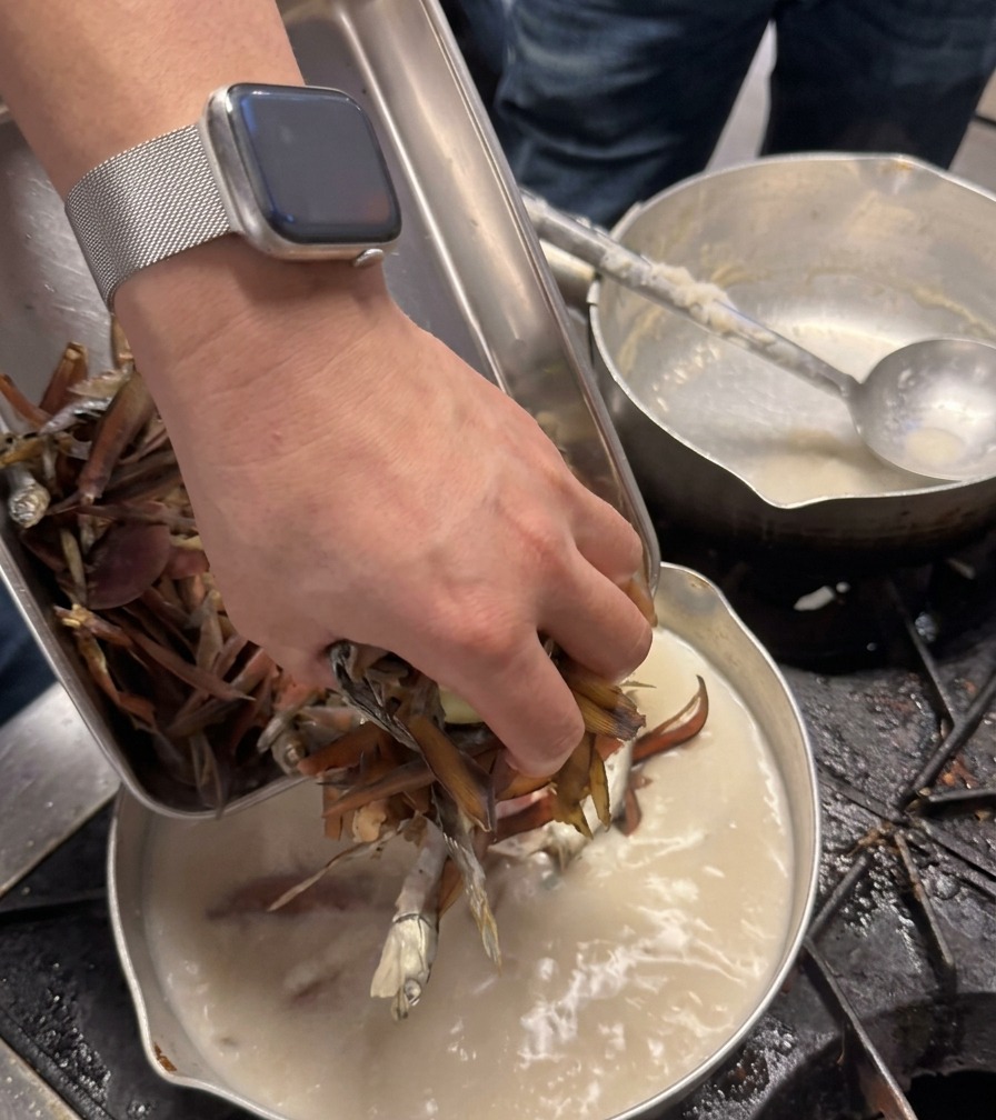셰프가 진하고 뽀얀 육수가 끓고 있는 냄비에 짙은 갈색의 말린 재료를 한 움큼 넣고 있는 손