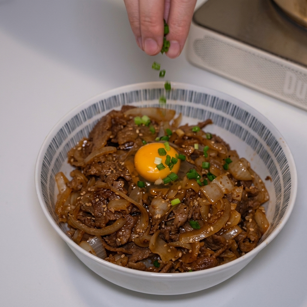 달걀노른자가 올라간 일본식 소고기 덮밥 위에 손가락으로 초록색 송송 썬 쪽파를 솔솔 뿌려 마무리하는 모습