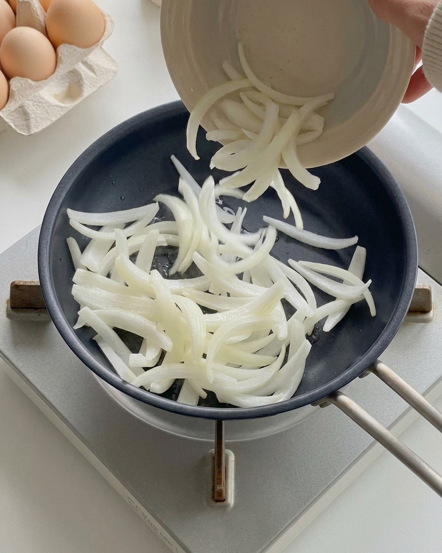 작은 볼에서 달궈진 검은색 프라이팬으로 채 썬 양파를 넣는 모습