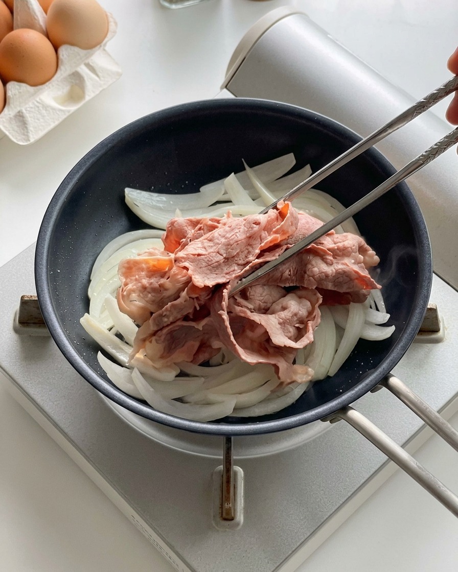 검은색 프라이팬에서 익어가고 있는 양파 위에 쇠젓가락으로 데친 소고기 슬라이스를 올리는 모습