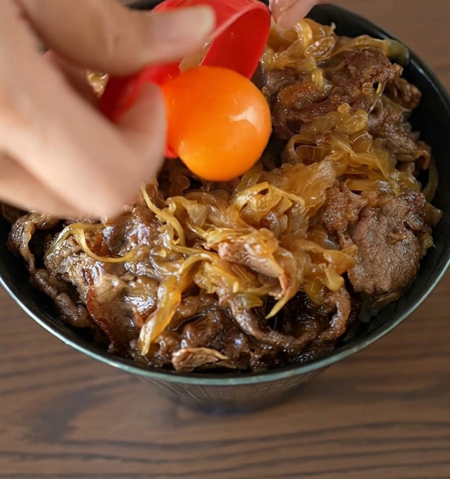 완성된 소고기 덮밥의 중앙에 빨간색 분리기에서 선명한 주황색 달걀노른자가 톡 떨어지는 모습.
