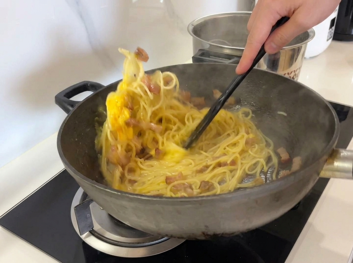 검은색 뒤집개를 이용해 웍 속의 스파게티와 계란 소스를 힘차게 섞고 있는 모습.