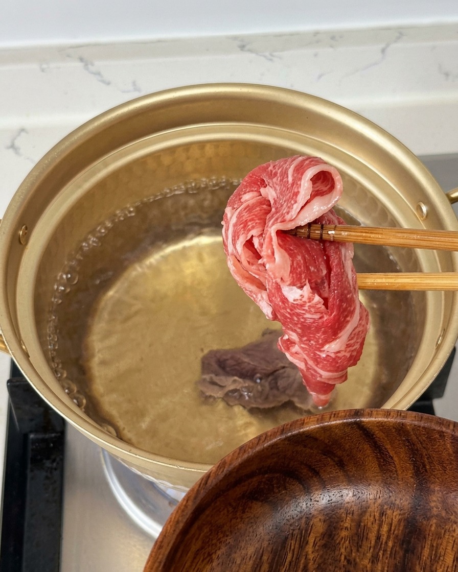 마블링이 있는 얇게 썬 생소고기 한 점을 젓가락으로 집어 끓는 물이 담긴 황금색 냄비 속으로 넣는 장면.