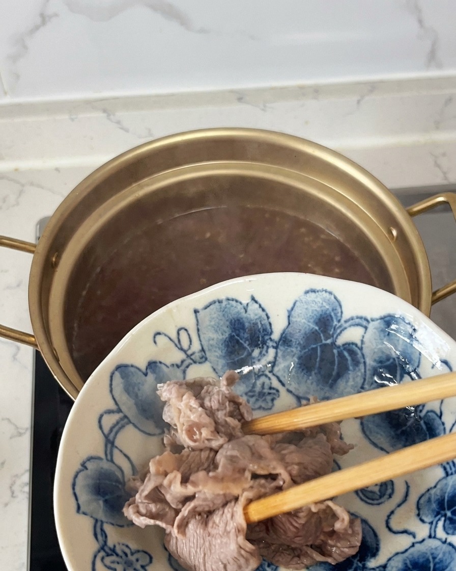 핏기가 가시고 살짝 익어 불투명해진 소고기 슬라이스를 젓가락으로 건져내어 파란 패턴이 있는 접시로 옮기는 모습.