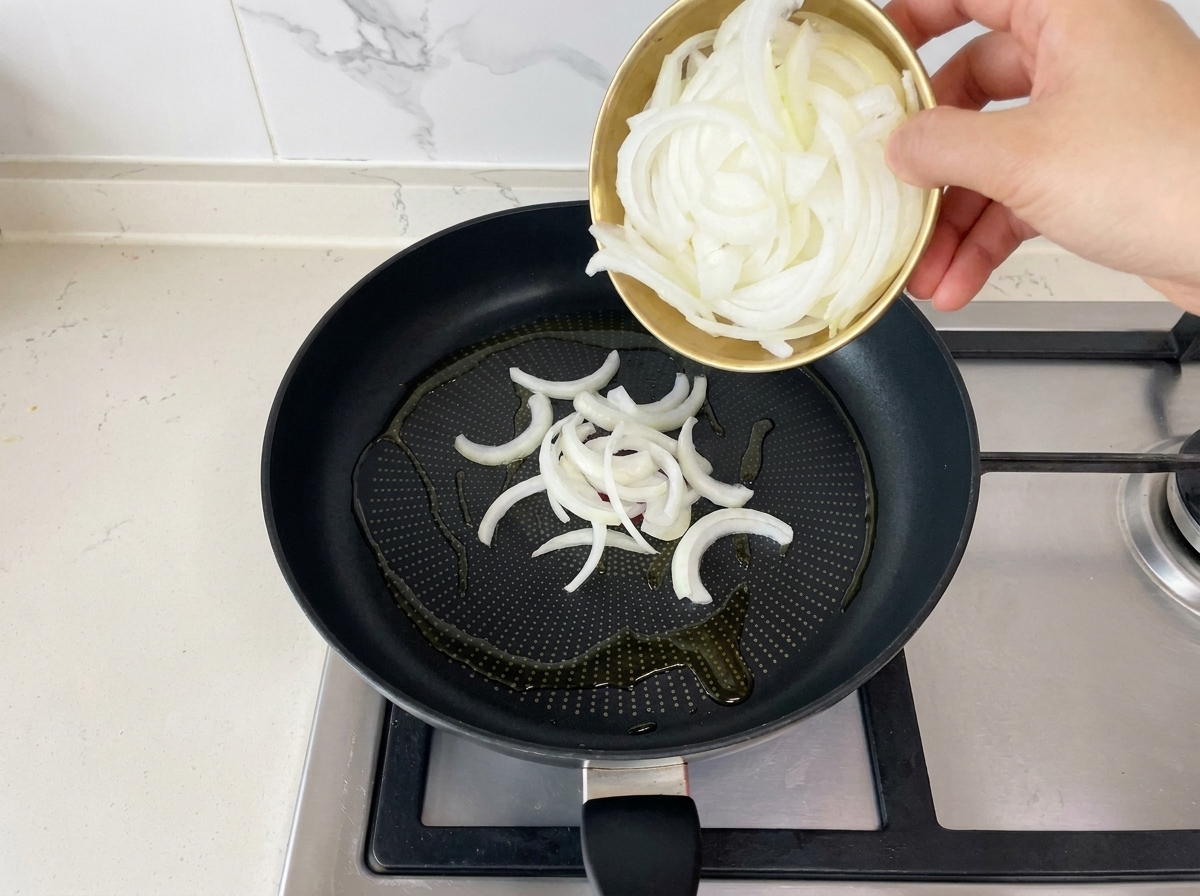 달궈진 기름이 두러진 검은색 프라이팬 위로 작은 황금색 볼에 담긴 채 썬 양파를 쏟아붓는 모습.
