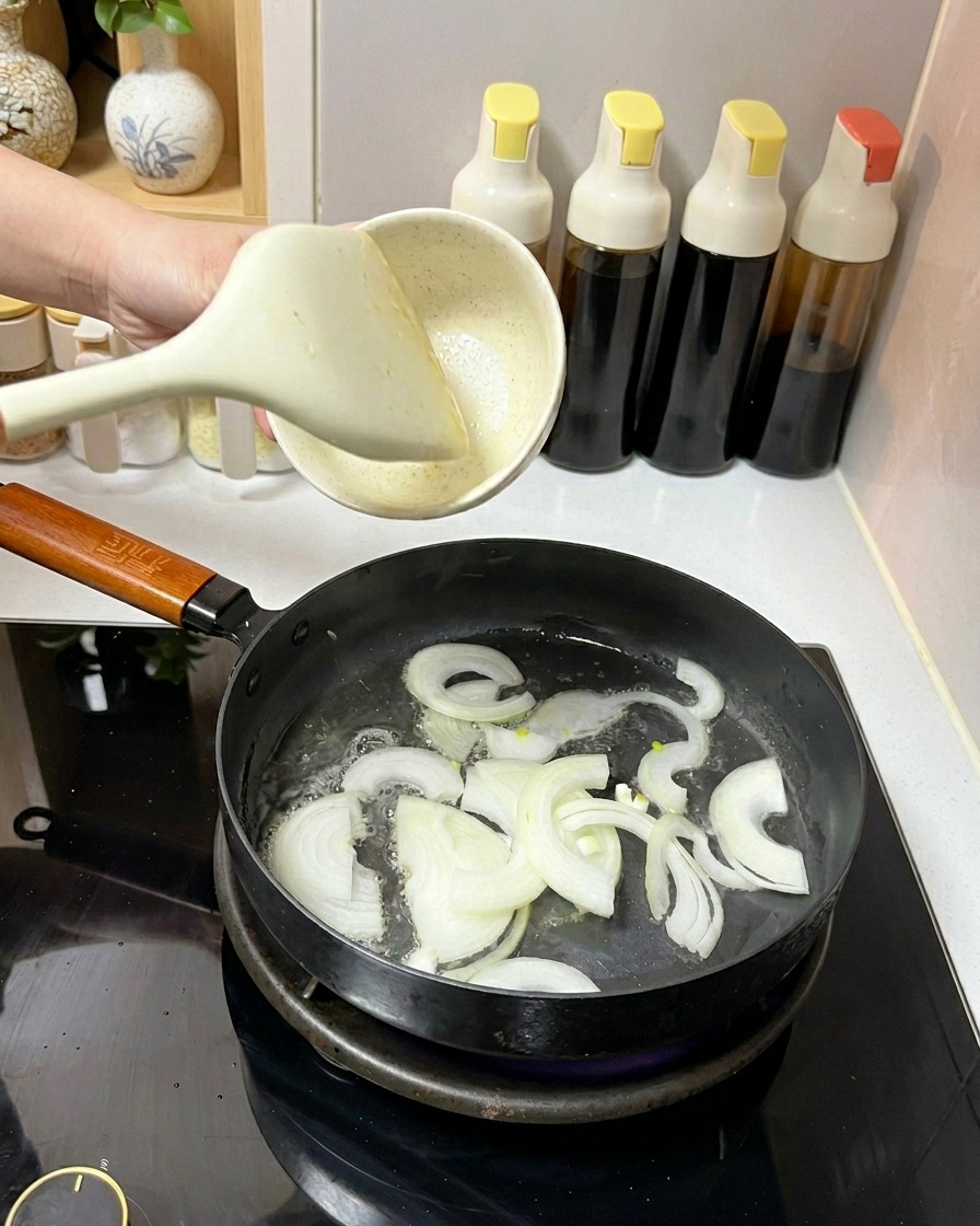 하얀 뒤집개를 사용해 기름을 두른 프라이팬에 채 썬 양파를 넣고 볶는 모습.