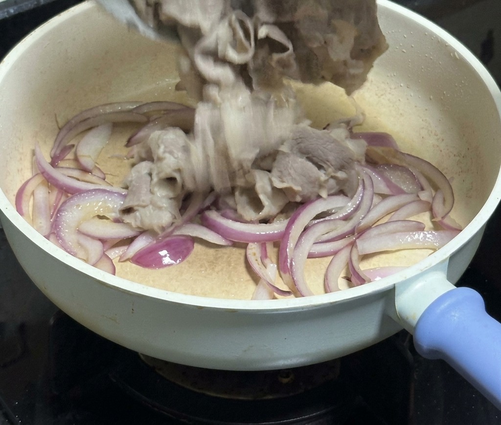 부드럽게 볶아진 적양파 위로 흰 볼에 담긴 데친 소고기를 프라이팬에 넣는 모습.