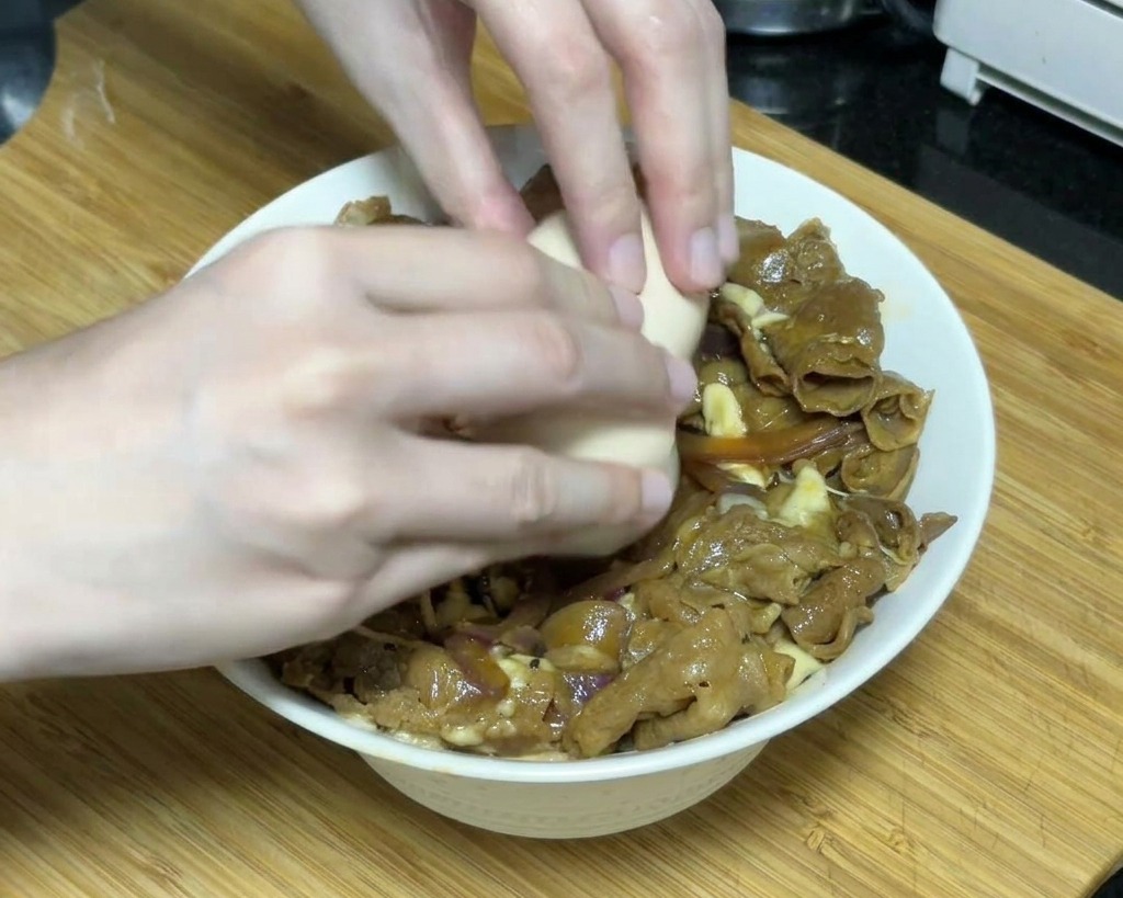 두 손으로 치즈가 녹아있는 데리야끼 규동 중앙에 부드럽게 익은 온센타마고를 조심스럽게 깨뜨려 올리는 모습.