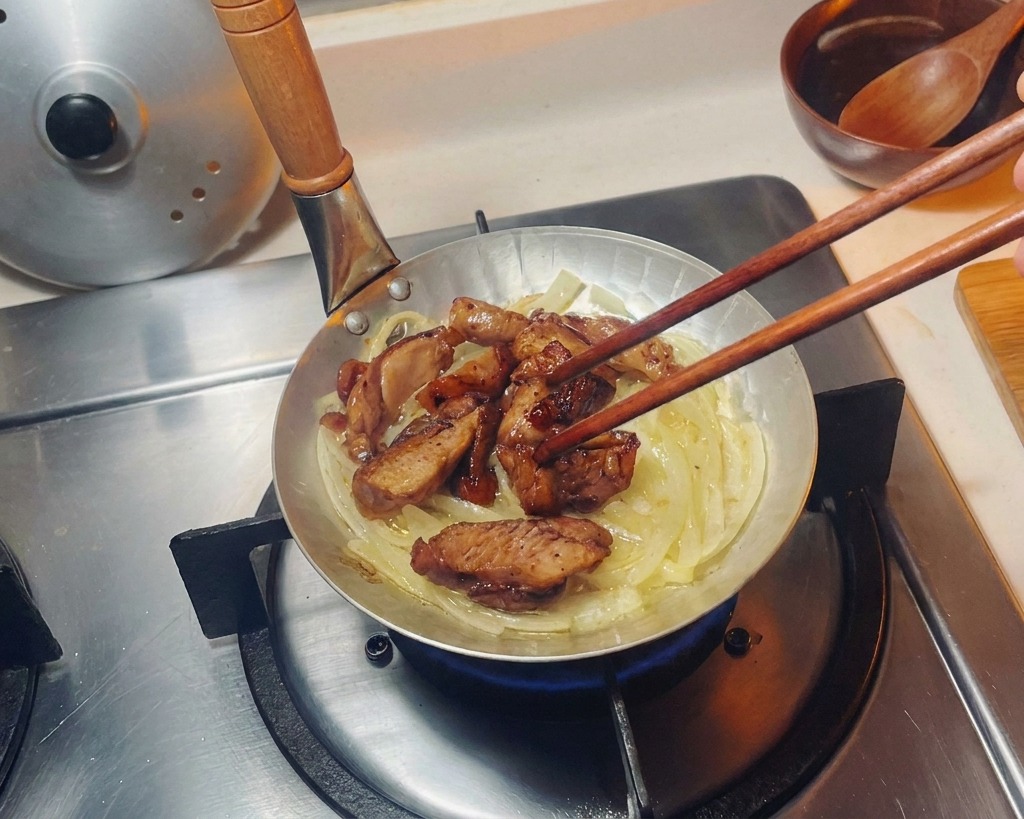작은 프라이팬 속 투명하게 익은 양파 침대 위에 젓가락으로 구운 닭고기 조각들을 가지런히 올리는 모습.