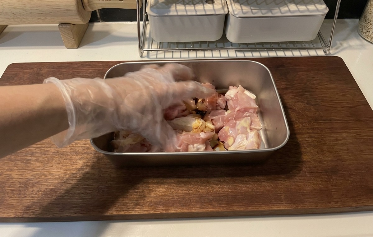 나무 도마 옆 금속 쟁반에서 위생 장갑을 낀 손으로 한입 크기의 닭고기에 간장 양념이 잘 배도록 버무리는 모습.