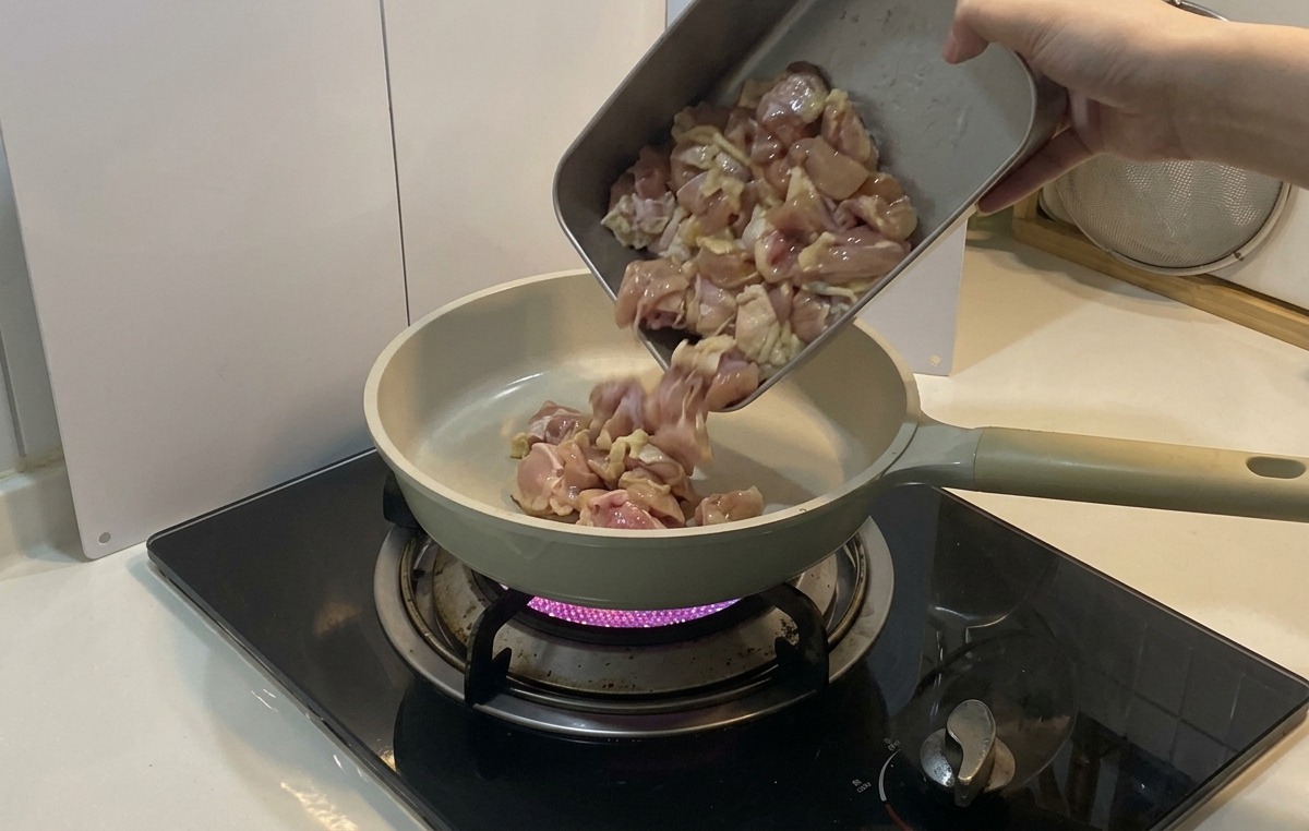 보라색 가스불 위에 올려진 밝은색 세라믹 프라이팬에 밑간한 닭고기 조각들을 금속 트레이에서 쏟아 넣는 모습.