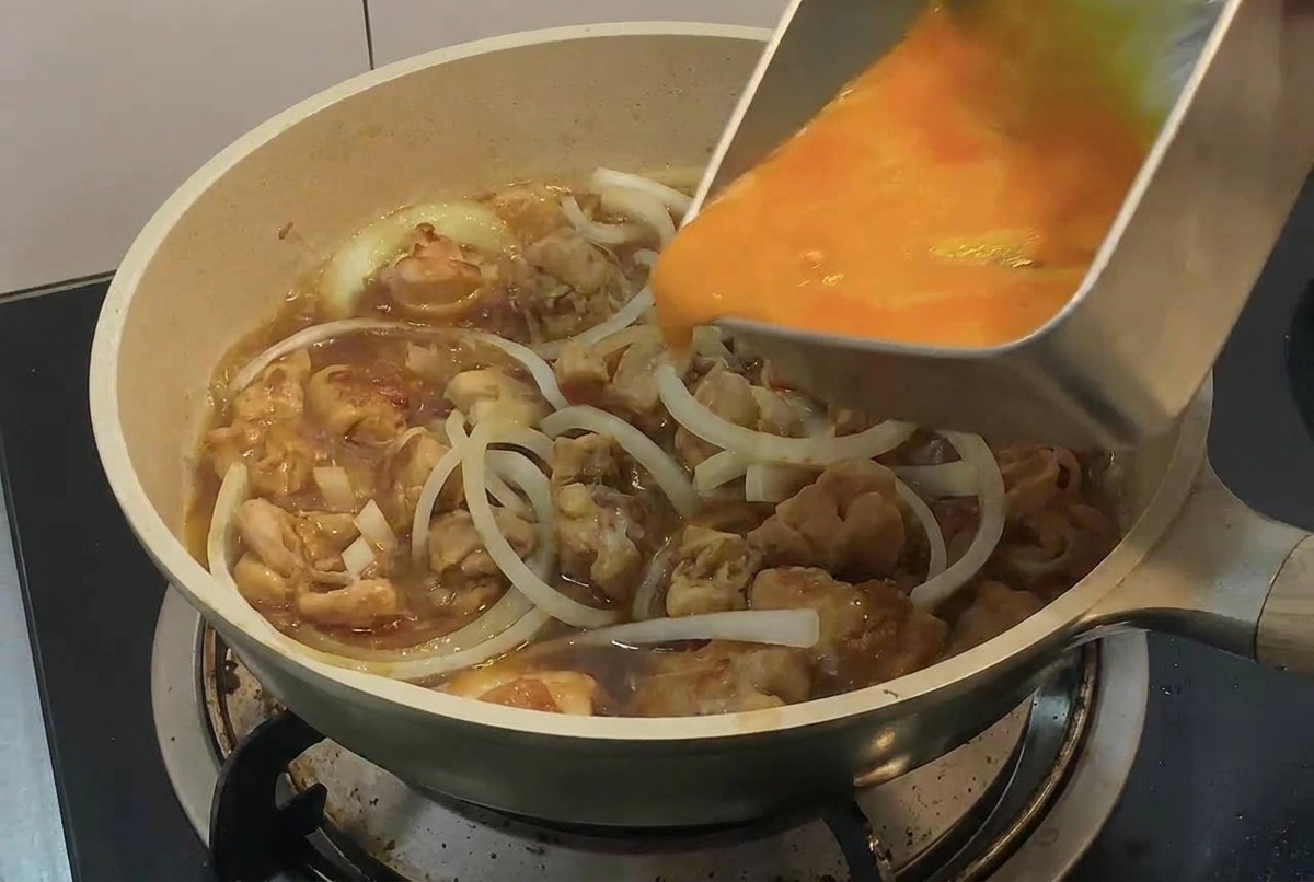 한 손으로 사각 금속 트레이를 들고 프라이팬 속 끓고 있는 닭고기와 양파 위로 대강 푼 달걀물을 둥글게 원을 그리며 붓는 모습.