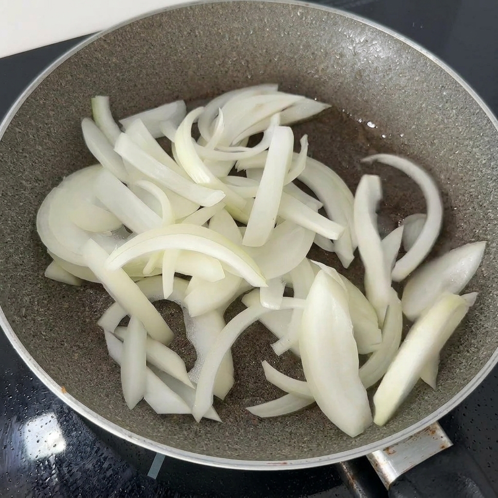 닭고기 기름이 남은 어두운 팬에서 채 썬 흰 양파가 부드러워질 때까지 볶아지는 모습