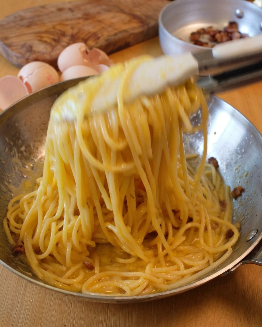 집게로 면을 공중으로 들어 올리며 크리미한 소스가 잘 섞이도록 팬을 흔드는 모습.