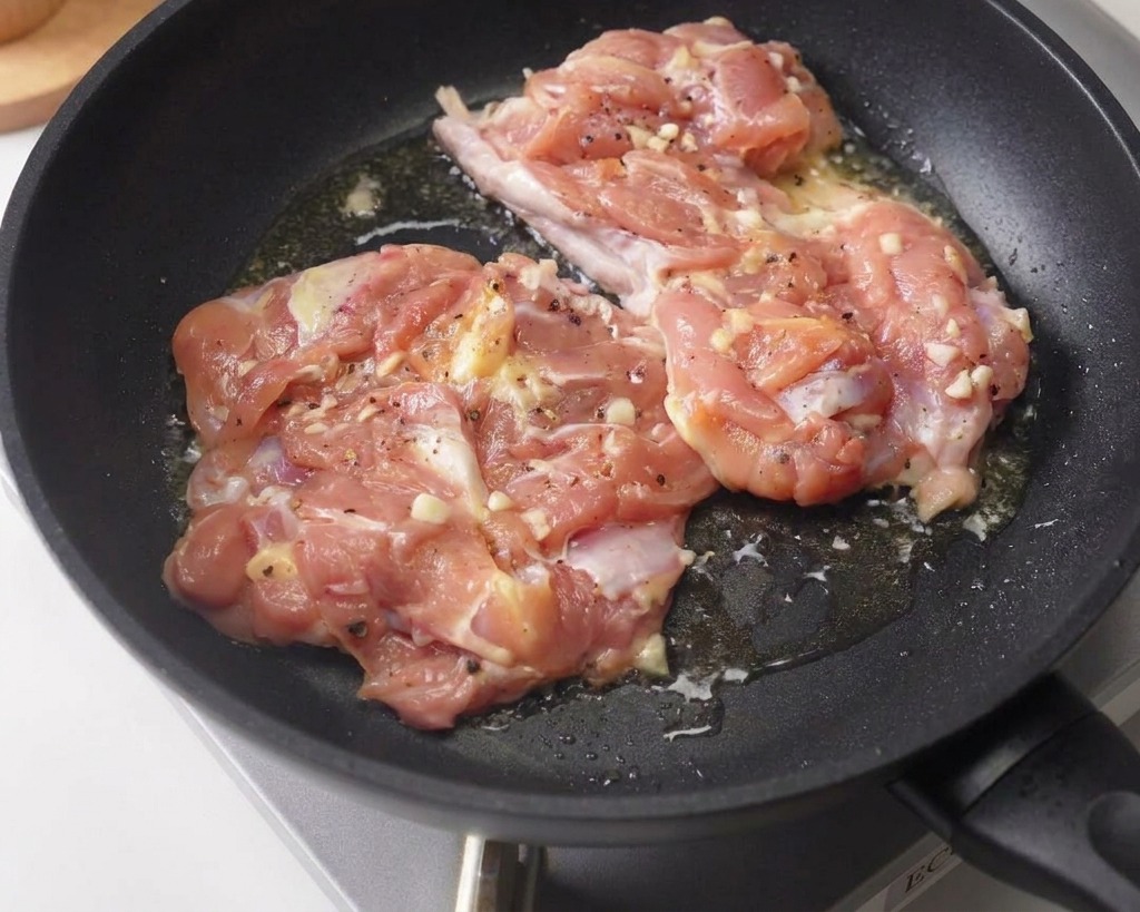 양념된 닭다리살 두 조각을 어두운색 프라이팬에 껍질이 아래로 가도록 올려 약불에서 굽는 모습.