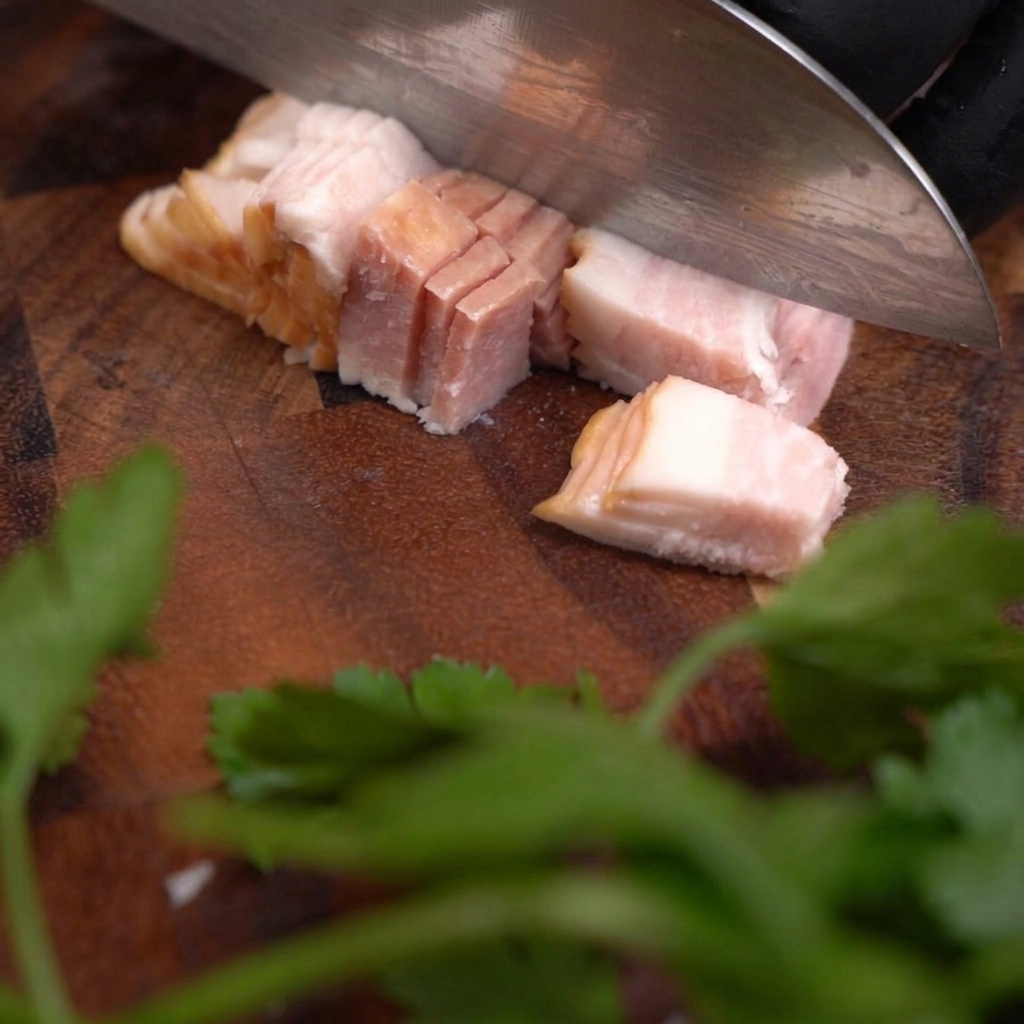 어두운 나무 도마 위에서 날카로운 칼로 생베이컨을 일정한 크기의 큐브 모양으로 써는 모습.
