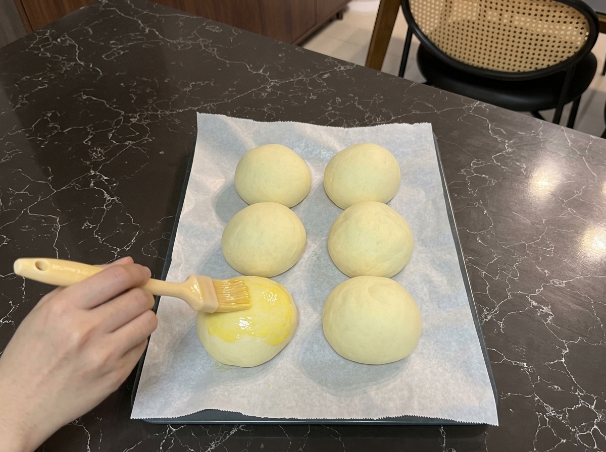 크게 부풀어 오른 발효된 버거 번 윗면에 페이스트리 붓으로 노란색 계란물을 부드럽게 바르는 모습.