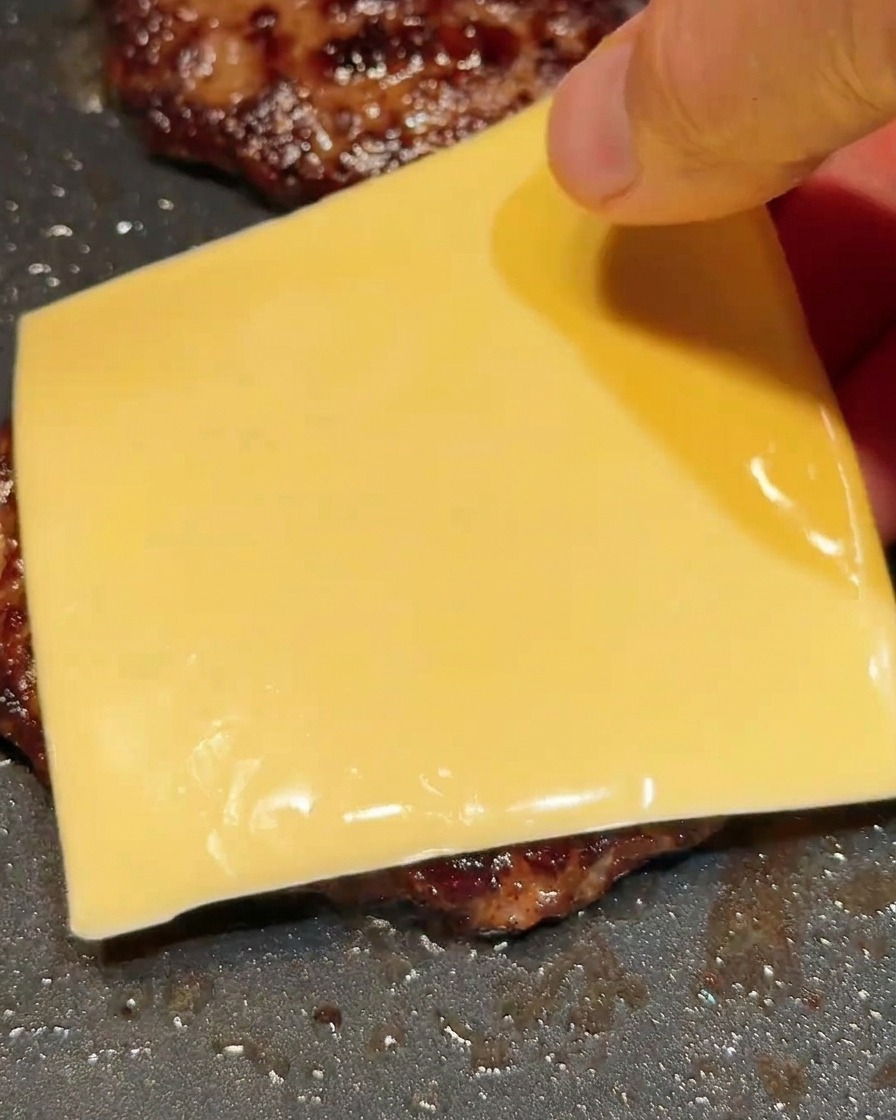 뜨거운 고기 패티 위에 노란 치즈 한 장을 올리는 모습.
