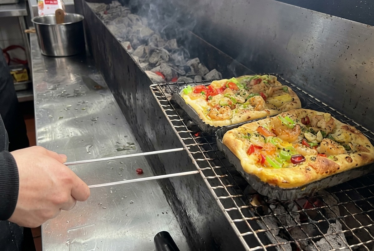 전문 주방에서 새우와 혼합 채소를 올린 사각형 피자를 긴 숯불 그릴 석쇠 위에 올리는 모습.