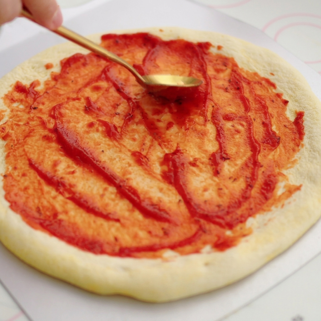 황금빛 스푼을 이용해 생 피자 도우 위에 진한 빨간색 토마토 소스를 원을 그리며 펴 바르는 모습