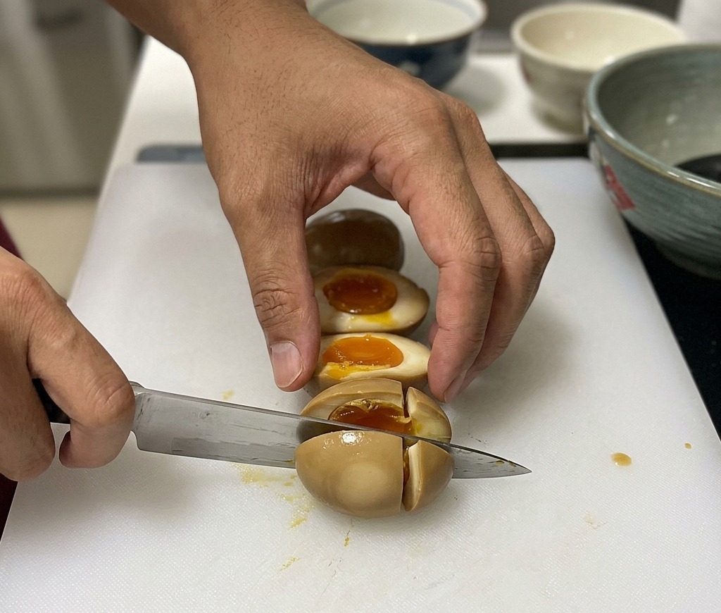 하얀 도마 위에서 셰프가 간장에 조려진 갈색 반숙란을 반으로 자르자 주황색 노른자가 흘러내리는 모습.